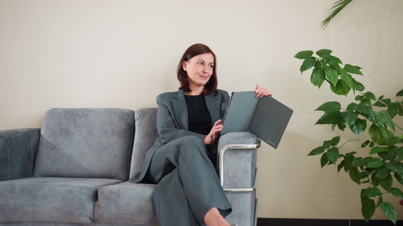 Confident businesswoman in gray suit relaxing on modern couch using digital tablet in cozy office setting with natural light and green indoor plant, enjoying remote work or online meeting