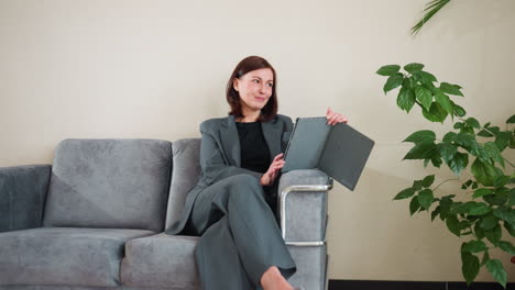 confident businesswoman in gray suit relaxing on modern couch using digital tablet in cozy office setting with natural light and green indoor plant, enjoying remote work or online meeting