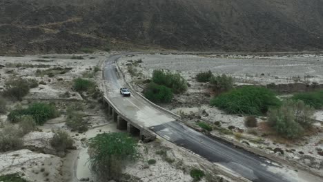 Toma-Aérea-De-Un-Automóvil-Blanco-Que-Viaja-En-Un-Puente-Bañado-Con-Agua-De-Río-Después-De-Una-Inundación-En-Baluchistán