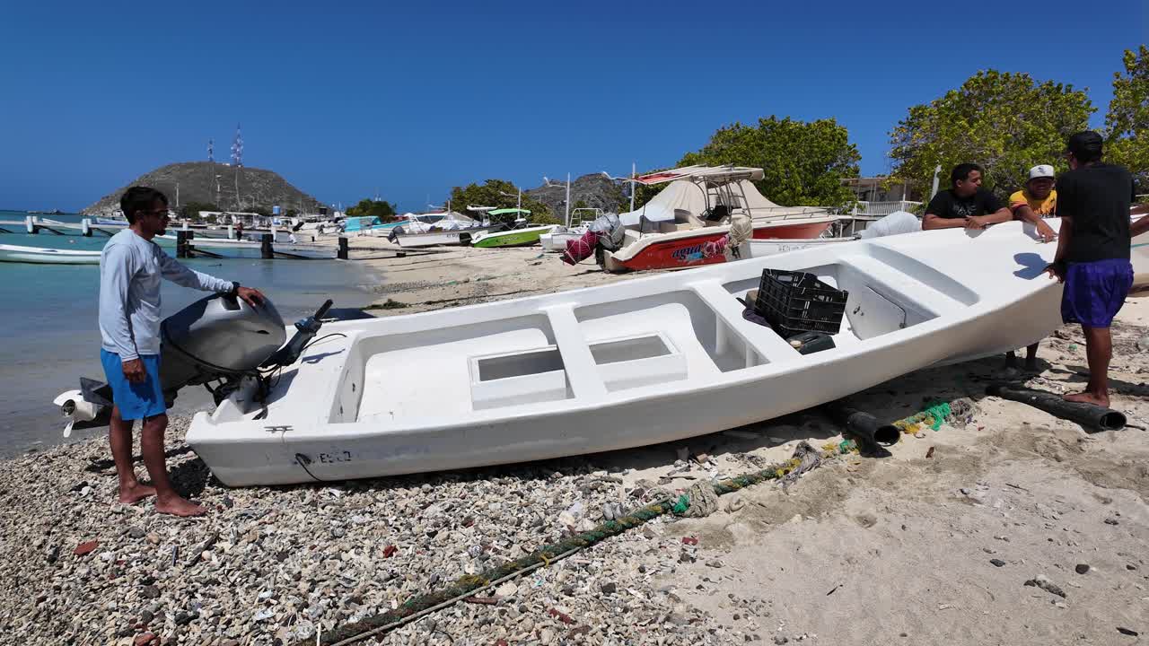 Scena costiera di uomini locali che preparano una piccola imbarcazione a motore su una spiaggia caraibica sabbiosa a Los Roques, Venezuela. Mezzi di sussistenza costieri, barche da pesca e imbarcazioni ancorate sono visibili sullo sfondo.
