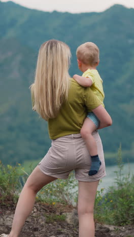 mother holds toddler boy in arms and points to river in valley against large mountains silhouettes at eco resort in summer backside view slow motion