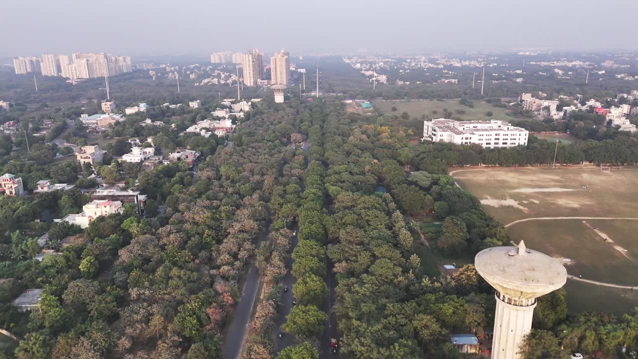 Riprese aeree con drone su Greater Noida mostrano edifici residenziali raggruppati che circondano un'area verde aperta, con strutture industriali in lontananza, in condizioni di lieve inquinamento atmosferico e foschia mattutina