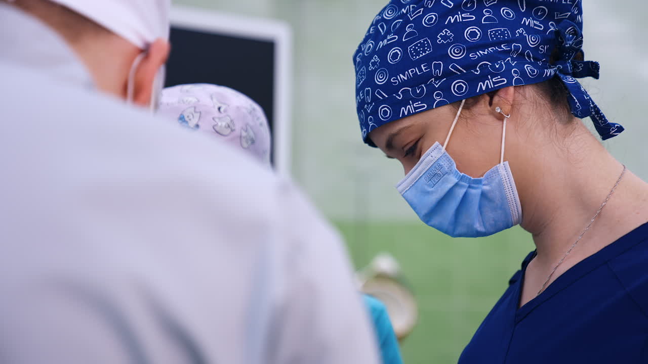 Female students in masks and caps smile and look down. Teacher showing something to the future medics.
