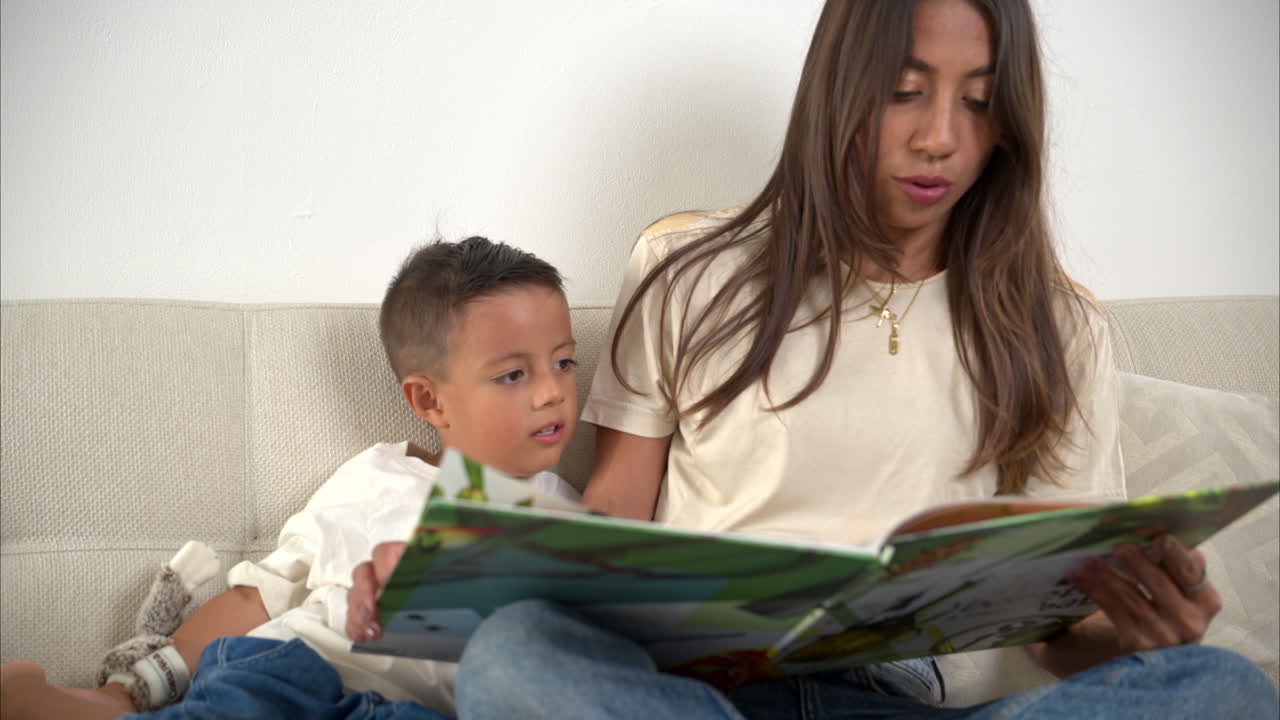 movimiento lento de un niño latino mexicano leyendo un libro con su madre sentado en el sofá con camisetas beige y vaqueros azules