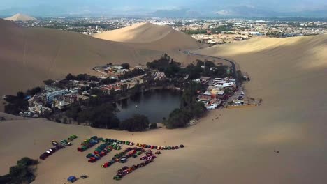 Aerial,-pull-back,-drone-shot,-rising-away-from-the-Huacachina-oasis-and-village,-on-a-sunny-day,-in-south-Peru