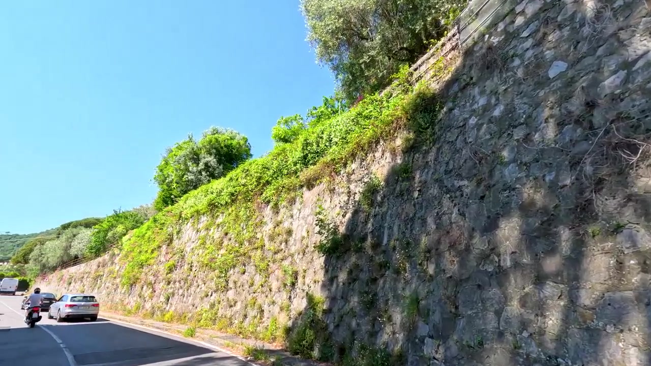 Prospettiva in prima persona mentre si guida lungo una strada mediterranea assolata, fiancheggiata da una vegetazione lussureggiante e muri in pietra.