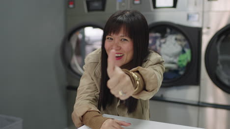 An-Asian-woman-with-bangs-exudes-immense-joy-and-excitement-making-a-thumbs-up-while-doing-her-laundry,-perhaps-celebrating-her-newfound-independence-or-a-significant-milestone-in-her-life