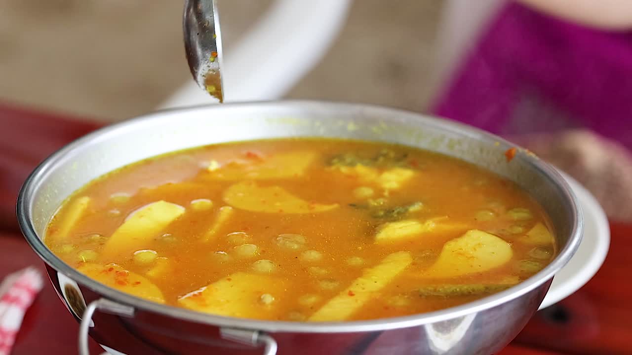 A ladle dips into a steaming pot of yellow curry, showcasing vibrant ingredients and rich broth.
