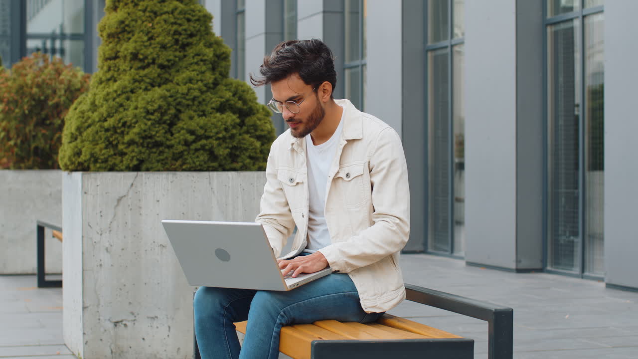 Indian man freelancer start working opens laptop sends messages reading email outdoors on bench