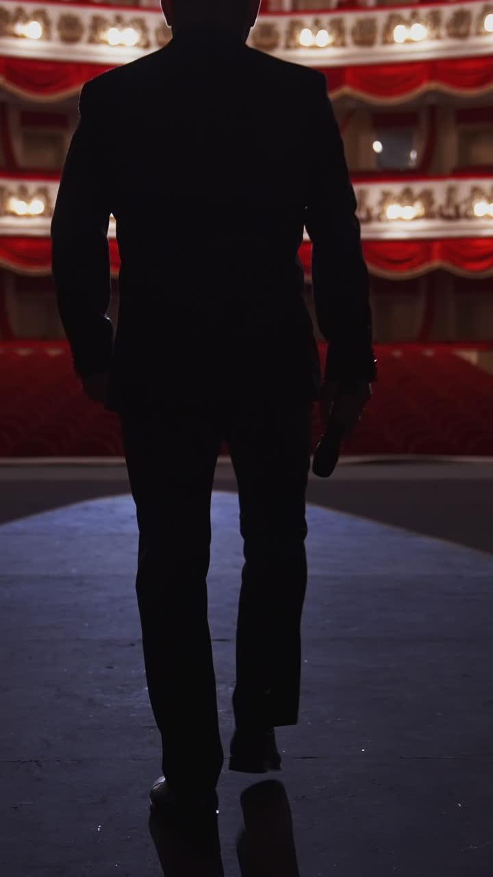 Man with microphone on theatre stage. Male actor in elegant suit walks on scene and starts speech in front of the empty auditorium. Back view. Vertical video