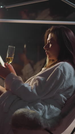 contemplative woman transfers glass between hands while sitting in comfortable chair. comfortable in dim light envelops pensive woman sitting in cozy robe