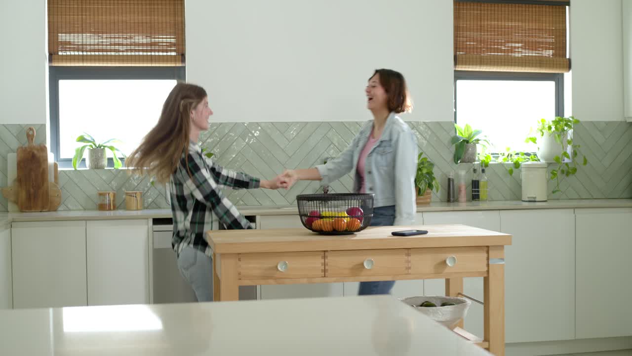 Mani che si cercano, due donne che girano e volteggiano intorno a un'isola da cucina in legno, mostrando affetto.
