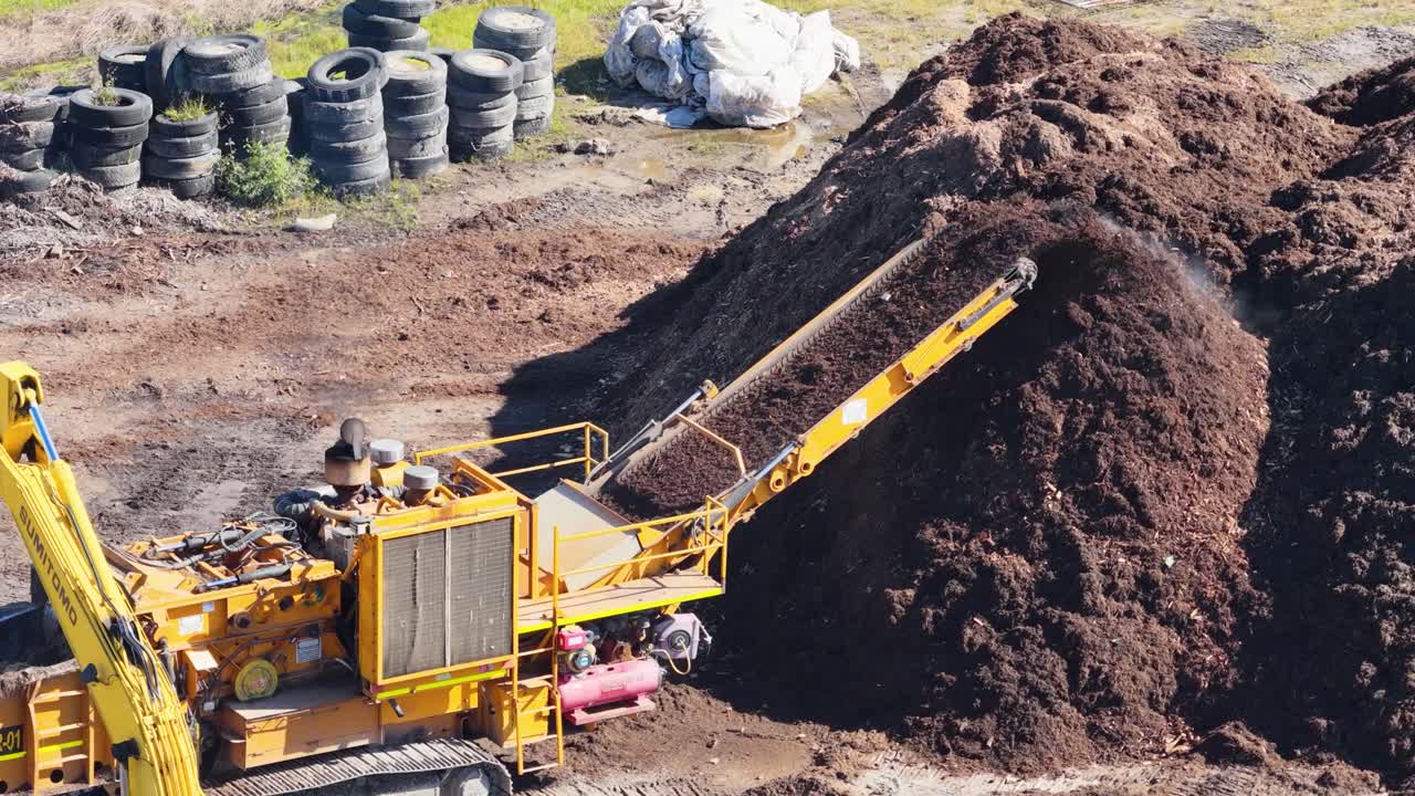 Drone footage captures an excavator moving mulch with precision. Bright daylight highlights the industrial process in a rural setting