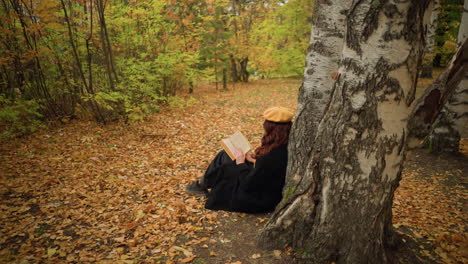 woman immersed in book, flipping through pages in peaceful solitude, leaning against tree in golden autumn forest, she enjoys literature and serenity, surrounded by fallen leaves and nature beauty