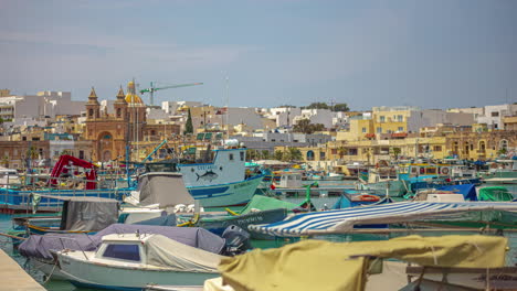 Zeitraffer-Von-Booten,-Die-Sich-Auf-Dem-Wasser-In-Der-Bucht-Von-Marsaxlokk,-Malta,-Bewegen,-Mit-Vielen-Gebäuden,-Häusern-Und-Einer-Kathedrale-Im-Hintergrund