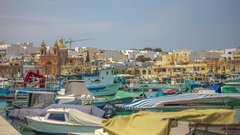 timelapse of boats moving around on the water in the bay of marsaxlokk, malta with many buildings, homes and a cathedral in the background