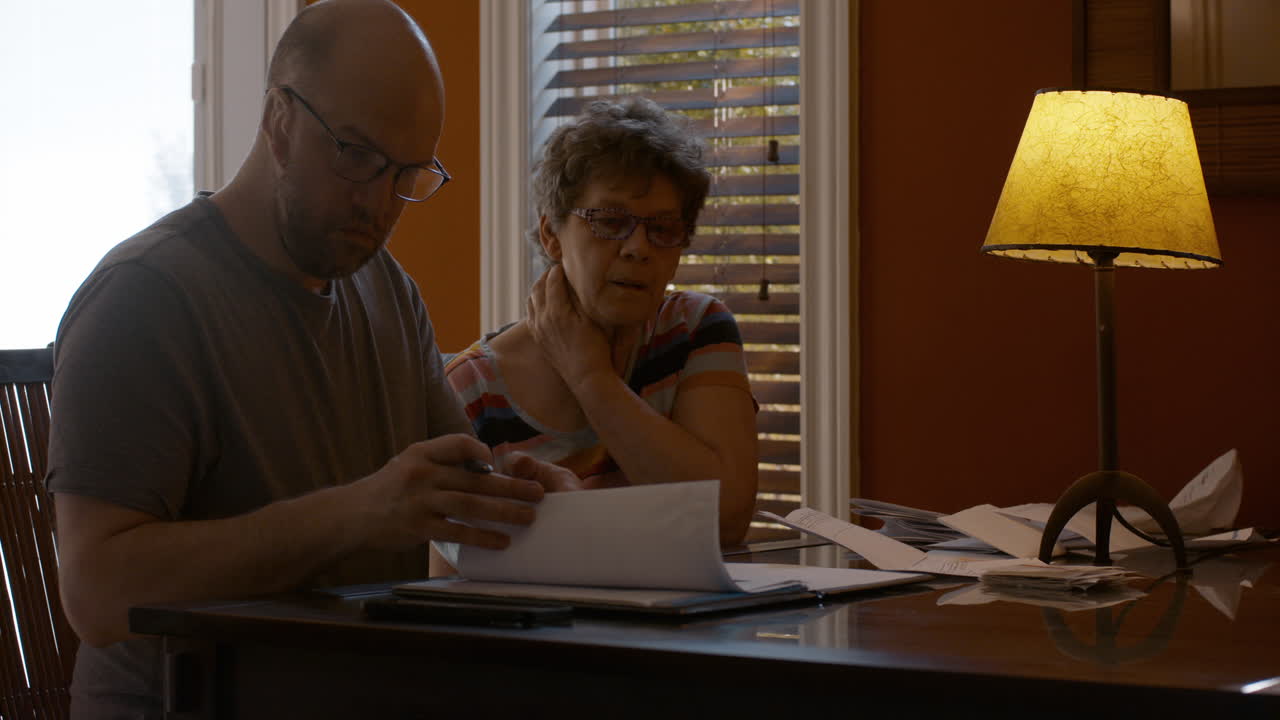 An adult son and his elderly mother sit together at a dining table in warmly lit home, sorting through paperwork. The son opens a notepad and gets organized, as mother is onlooking, bored.
