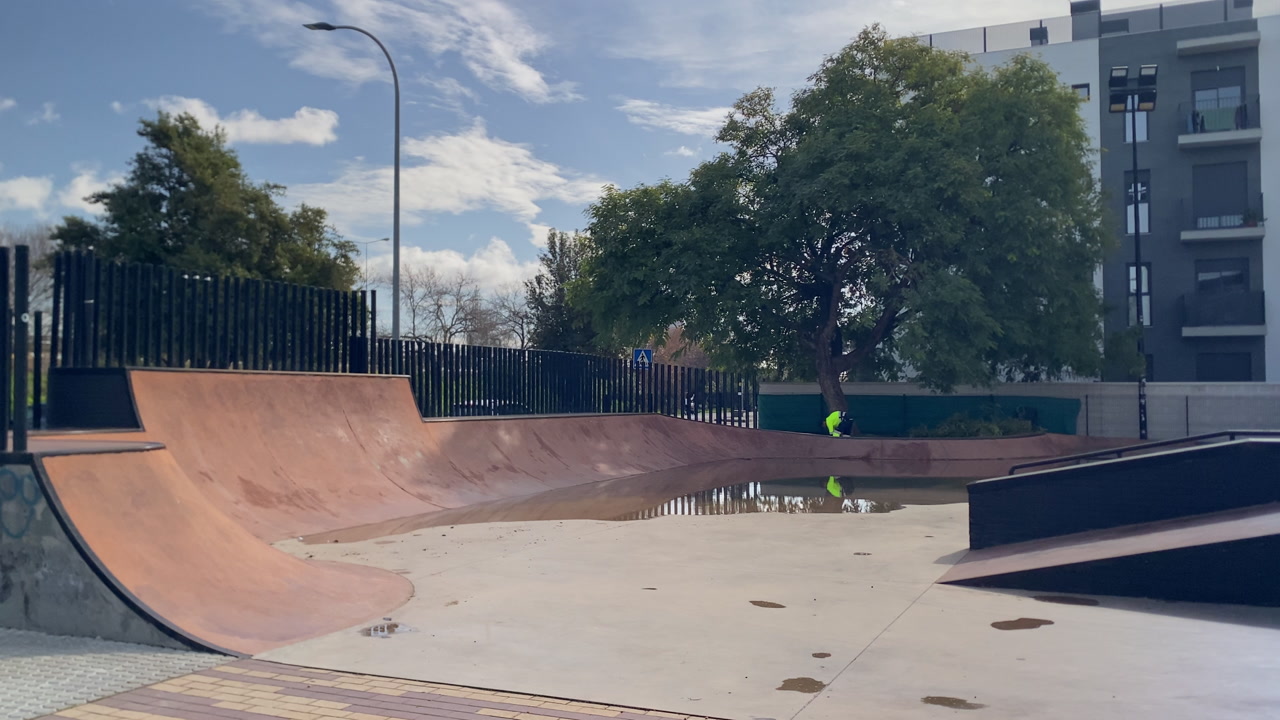 Urban skatepark bowl filled with rainwater, reflecting a person in a high visibility vest near a green tree under a cloudy sky