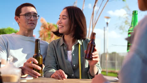 Gruppe-Von-Drei-Glücklichen-Japanischen-Freunden,-Die-Mit-Bier-Anstoßen,-Während-Sie-An-Einem-Sonnigen-Tag-Draußen-Am-Tisch-Sitzen