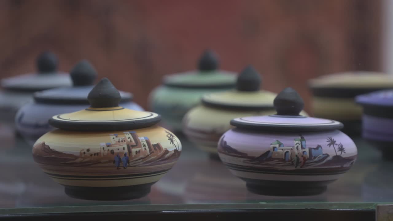 Close-up shot of traditional handmade ceramic jars displayed in a shop. The pottery features hand-painted Moroccan landscapes and colorful glazes in the souk