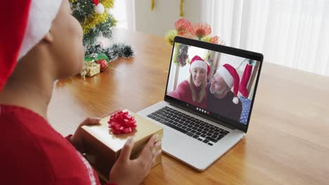 Afroamerikanische-Frau-Mit-Weihnachtsmütze-Nutzt-Laptop-Für-Weihnachtsvideoanruf,-Mit-Paar-Auf-Dem-Bildschirm