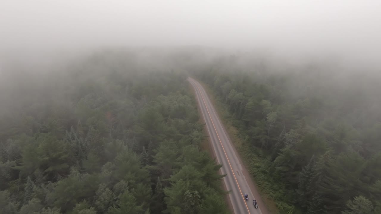 Drone following 2 motorcyclists winding through Northern Ontario forest through cloud cover