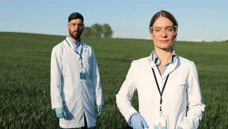 investigadores caucásicos con bata blanca, máscara y gafas mirando la cámara en el campo verde