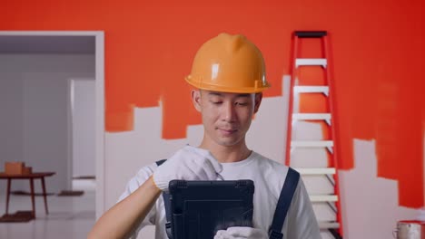 close up de un pintor asiático que usa un casco de seguridad tomando nota en una tableta mientras está de pie con la pared de pintura en la habitación