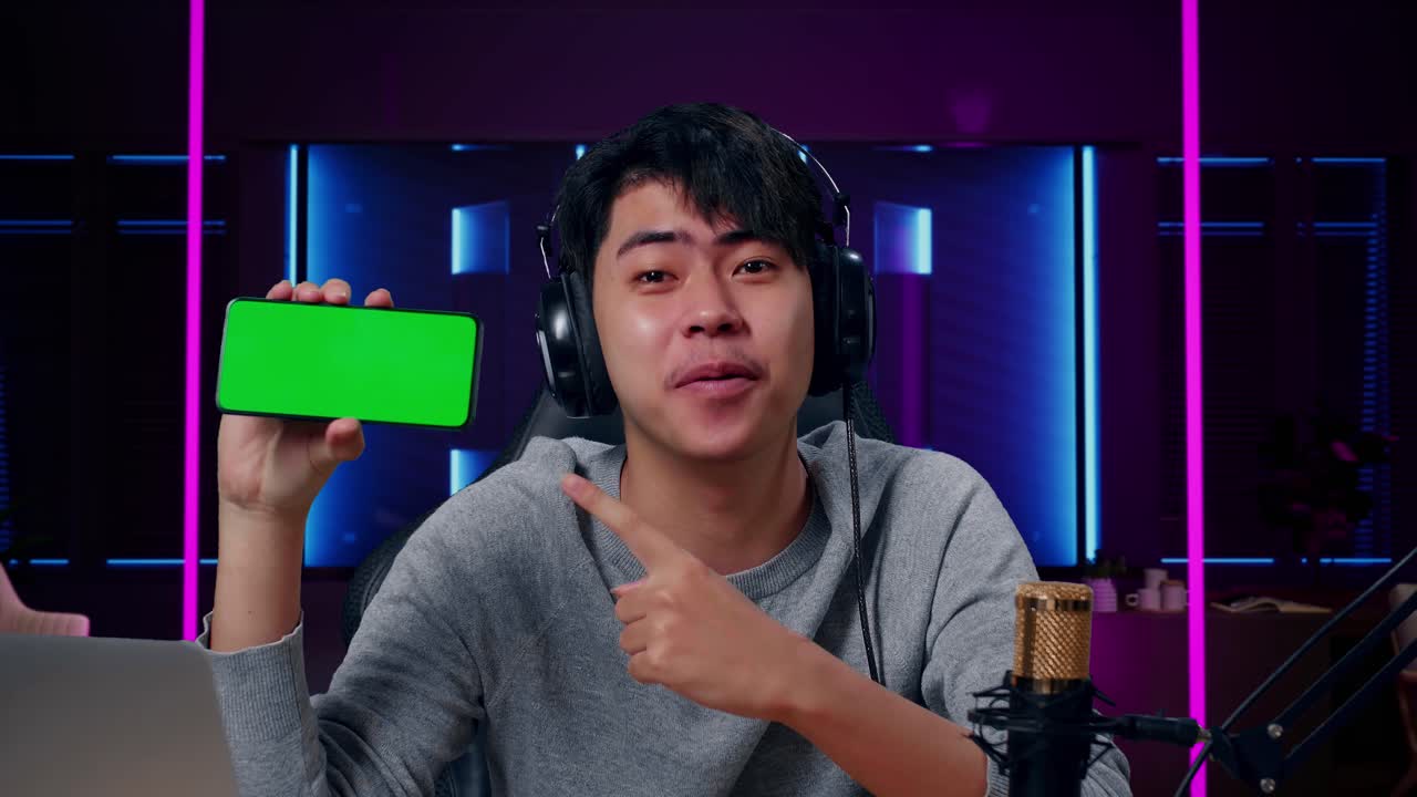 헤드폰과 컴퓨터를 들고 미소 짓는 아시아 남자의 근접 사진. 녹색 화면의 휴대전화가 레트로 네온 조명으로 조명된 방에 있습니다.