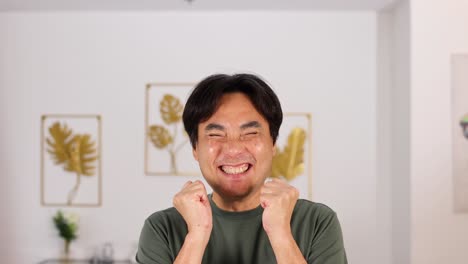 an asian man transitions from a surprised gesture to a joyful, smiling expression in a brightly lit, modern living room with framed botanical art