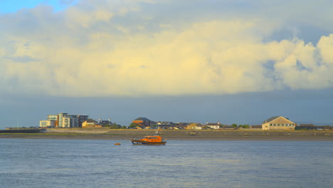 Stormy-sky-sweeping-by-and-rainbow-appearing-with-rain-bands-on-horizon,-as-moored-lifeboat-moves-with-incoming-tide-at-river-estuary-during-sunset