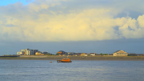 Ein-Stürmischer-Himmel-Zieht-Vorbei-Und-Ein-Regenbogen-Erscheint-Mit-Regenbändern-Am-Horizont,-Während-Sich-Das-Festgemachte-Rettungsboot-Bei-Sonnenuntergang-Mit-Der-Flut-An-Der-Flussmündung-Bewegt