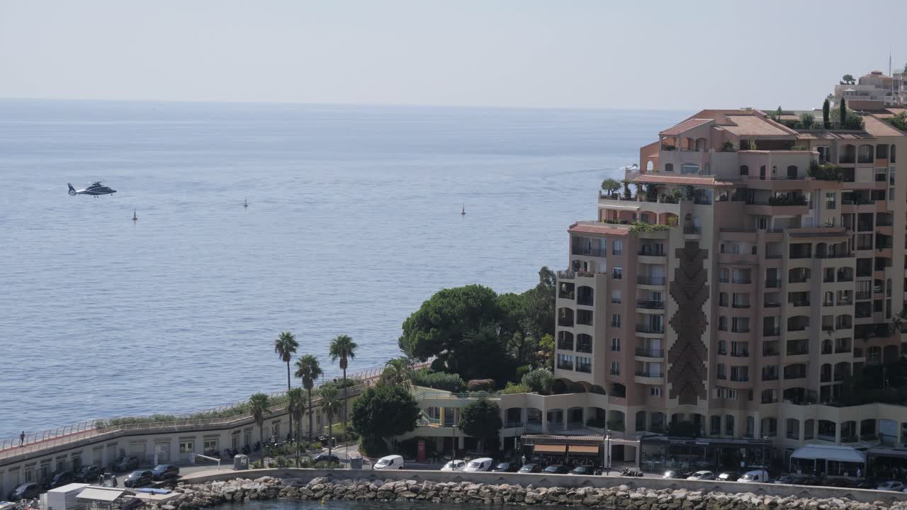 Un elicottero si avvicina a una piattaforma di atterraggio dietro a lussuosi condomini a Monaco. Il panorama sul lungomare e l'orizzonte mediterraneo creano una prestigiosa scena di viaggio e trasporto.