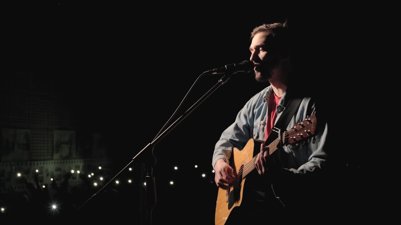 A musician is playing guitar on a large stage with audience members holding flashlights in the background. The auditorium is filled with beautiful flashing lights.