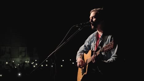 a musician is playing guitar on a large stage with audience members holding flashlights in the background. the auditorium is filled with beautiful flashing lights.