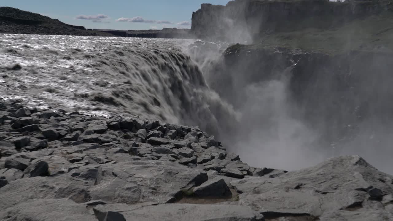 Zoom out shot of strong Dettifoss Waterfall crashing down rocky edge during sunny day in Iceland