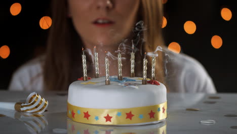 close up of a woman blowing out seven lit candles on a white, decorated birthday cake, a party blower beside it, bokeh lights in the background