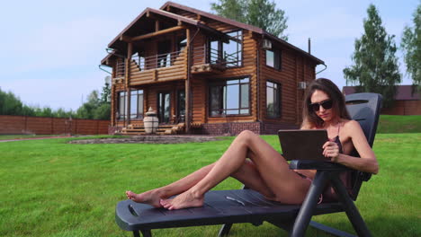 una hermosa mujer delgada se acuesta en una tumbona en un traje de baño con una tableta en el fondo de una gran casa de campo en gafas de sol y trabaja de forma remota en vacaciones