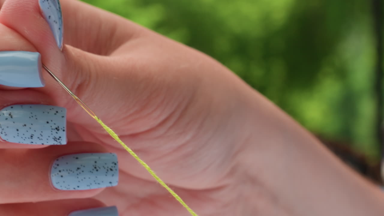 Close up hands with blue nails threading needle outdoors, fine green strand aimed into eye, careful craft prep in park, soft daylight, shallow bokeh, patient motion before embroidery work