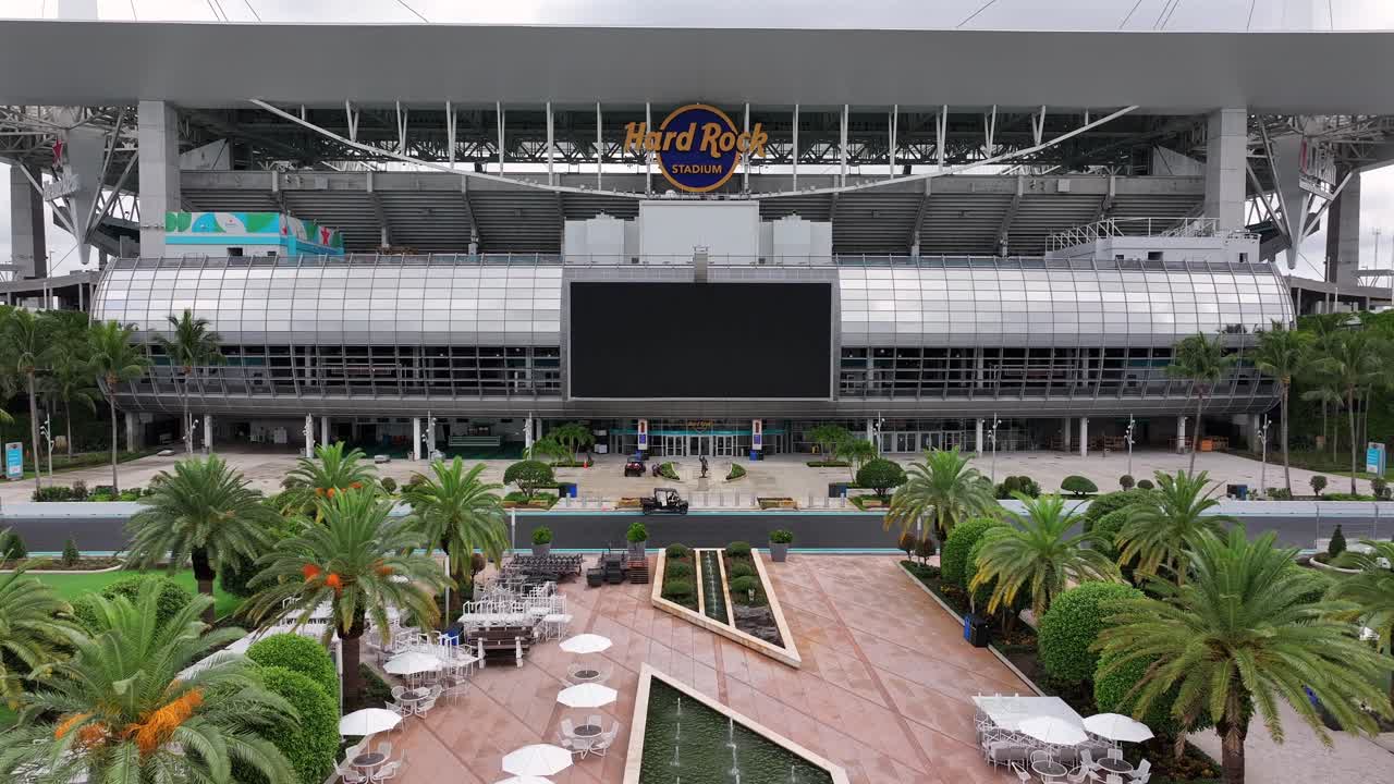Hard Rock Stadium in Miami, featuring its iconic roof canopy and large digital screen. The shot focuses on the stadium’s exterior with structural details visible. Aerial.