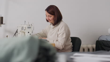 smiling woman carefully sewing fabric in bright workspace, radiating satisfaction, enjoying creative process, highlighting skillful hands, comfortable posture, and focus on garment creation