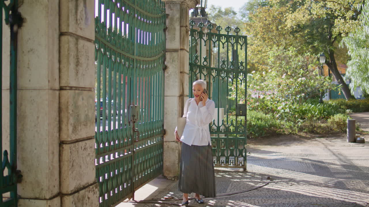Grey hair woman calling mobile phone waiting friend in park view through gate.