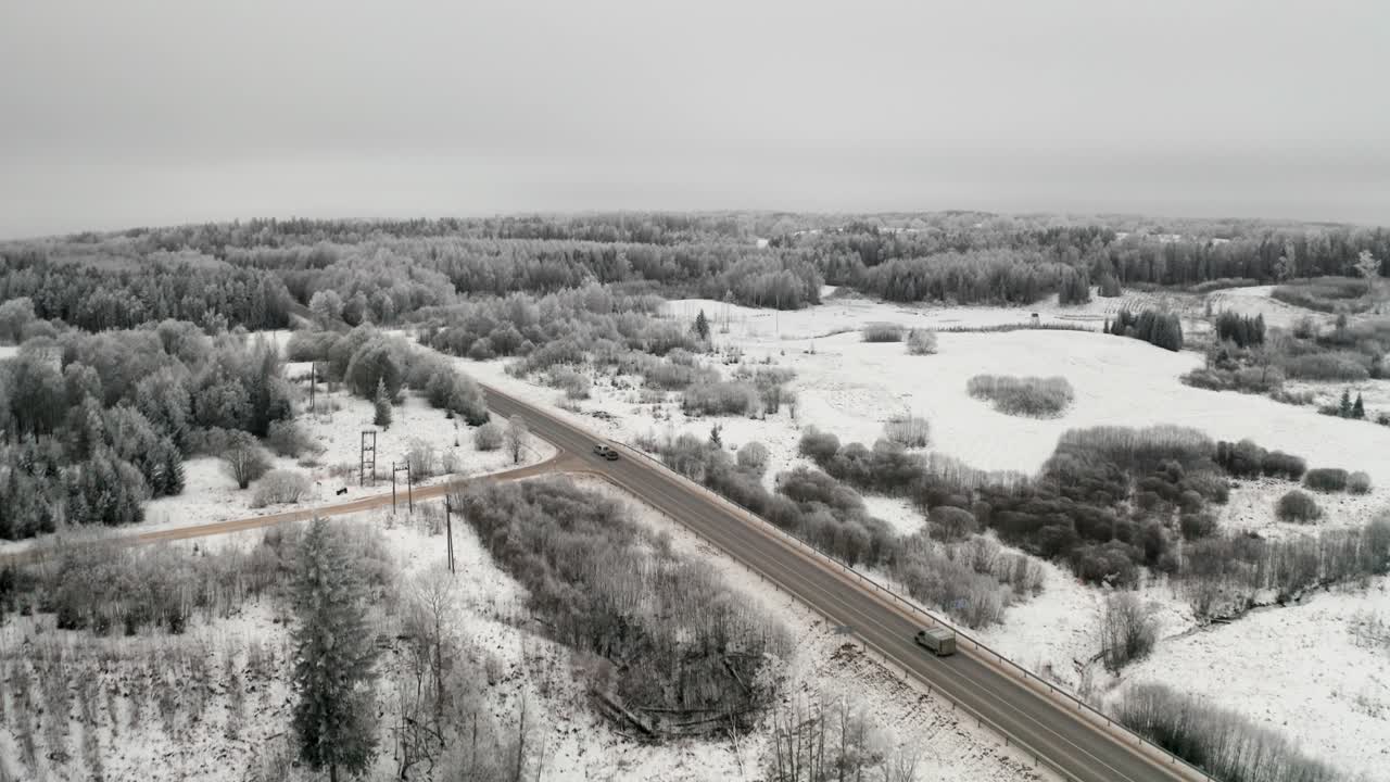 Aerial view of a two lane highway in countryside with cars passing by. Asphalt road intersection with gravel road. Frost over the landscape in winter season.