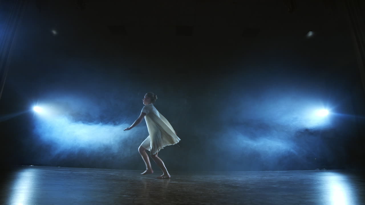 The zoom camera moves the woman dancer across the stage with software and smoke. A modern dramatic ballet a woman in a white dress spins on one leg and jumps..