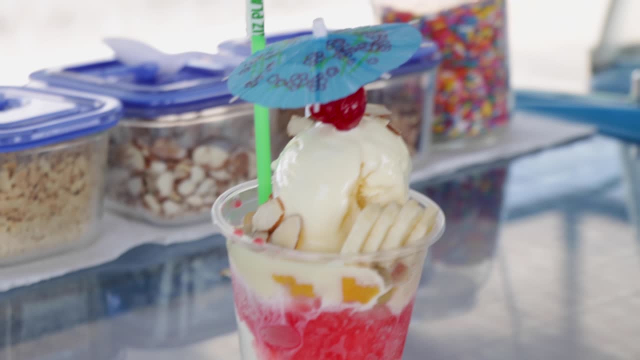 cono de nieve con hielo afeitado, jarabe de azúcar, helado, fruta de plátano y cereza en la parte superior, postre de playa servido en una taza de plástico, con contenedores de tapa en el fondo, costa rica