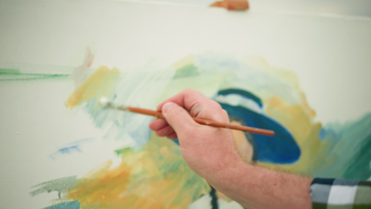 A close-up of an artist's hand, clad in a checkered shirt, as it skillfully paints a colorful portrait on a canvas. The scene captures the artist s meticulous brushwork, bringing the artwork to life