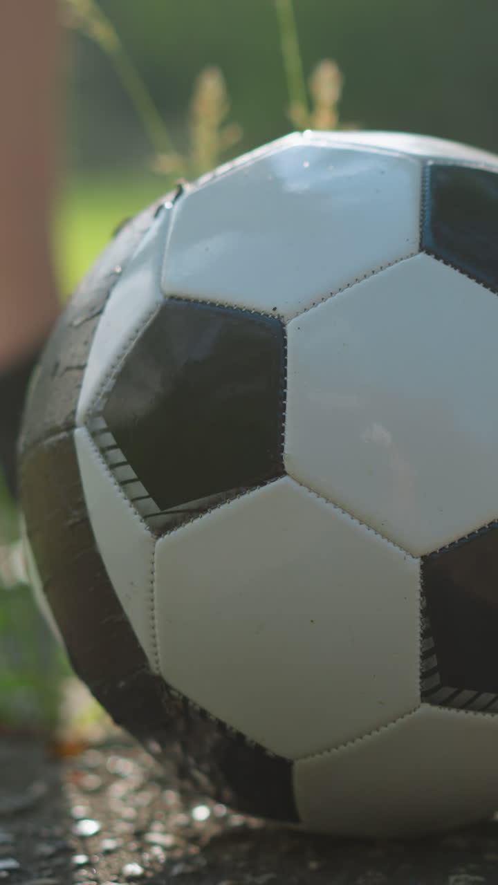 A close-up view of a soccer ball resting on a paved surface, with a blurred figure standing nearby, the background features a grassy field
