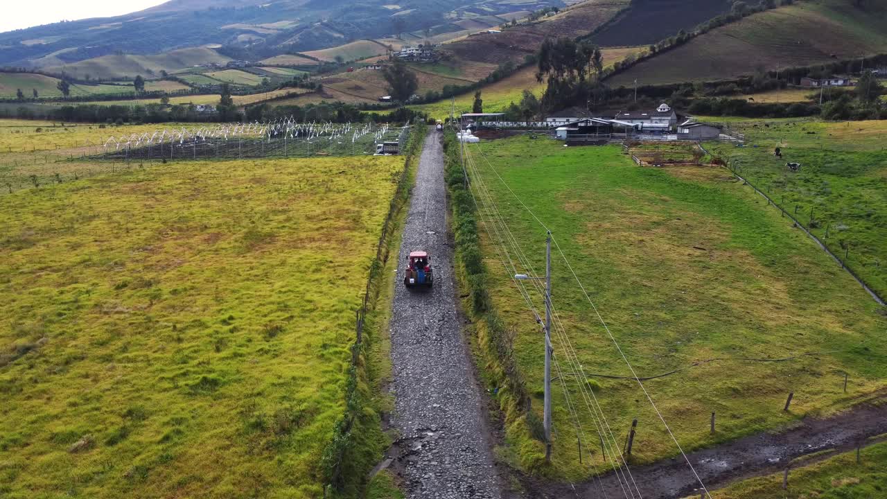 Aerial perspective capturing a tractor traversing a gravel road between vast, lush fields, showcasing the serene and expansive rural landscape with rolling hills in the background under a clear sky