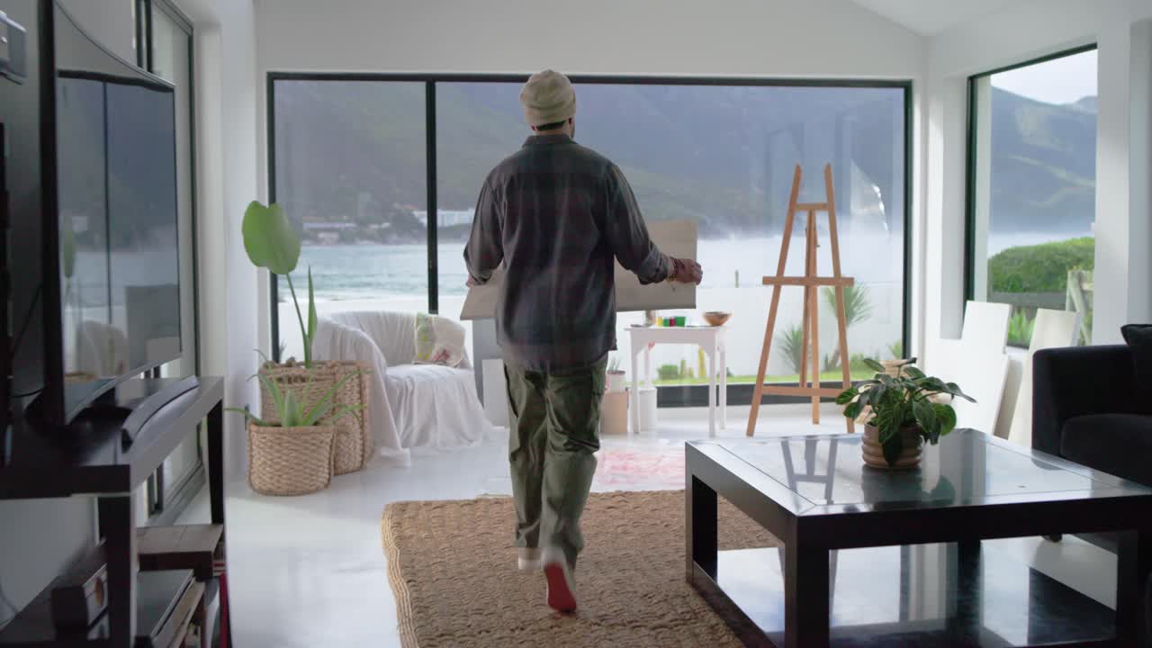 Man is entering doorway, carrying canvas toward easel by window, mounting and preparing to paint