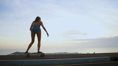 Captured-in-slow-motion,-a-stunning-young-skateboarder-rides-her-board-in-shorts-along-a-mountain-road-at-sunset,-with-the-mountains'-breathtaking-view-enhancing-the-scene